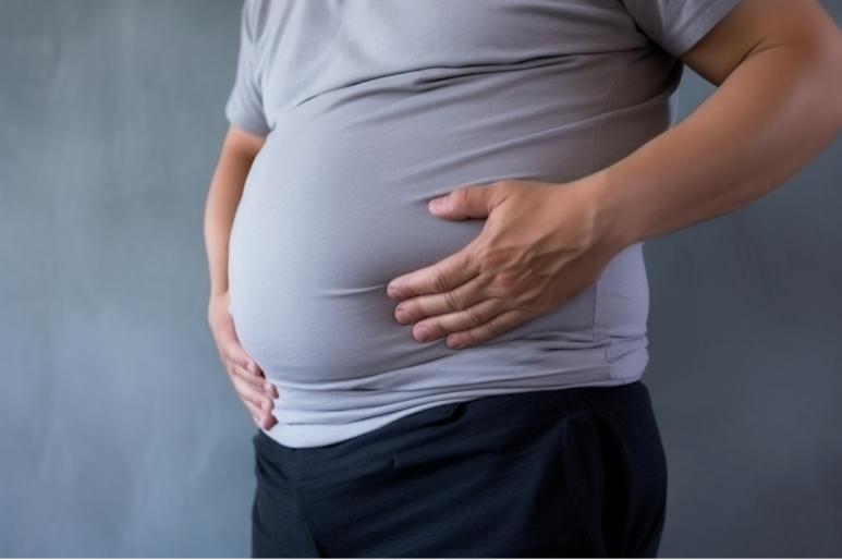 회색 티셔츠와 검은 바지를 입은 남성이 배를 두 손으로 잡고 있는 모습. 배가 앞으로 불룩하게 나와 있으며, 배경은 회색 벽으로 단순하게 구성되어 있다