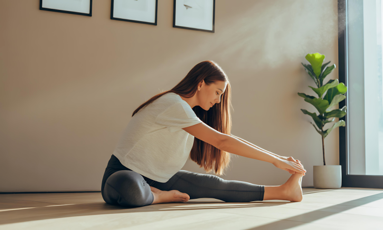 밝은 햇살이 드는 방에서 여성이 바닥에 앉아 스트레칭을 하는 모습. 한쪽 다리를 곧게 뻗고 손으로 발끝을 잡으며 상체를 숙이고 있다. 그녀는 흰색 티셔츠와 회색 레깅스를 입고 있으며, 옆에는 화분이 놓여 있다