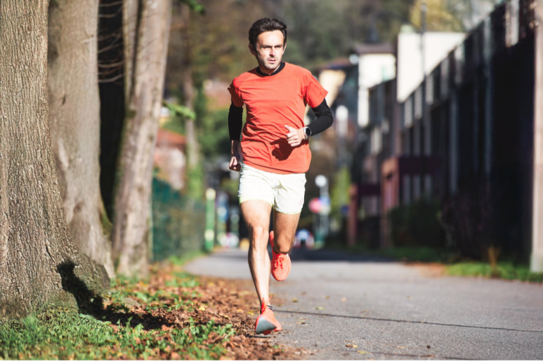 낮 동안 햇살이 비치는 거리에서 달리기를 하는 남자의 모습. 그는 주황색 티셔츠, 흰색 반바지, 주황색 운동화를 착용하고 있으며, 왼쪽 팔목에는 검은색 웨어러블 기기를 착용하고 있다. 그가 달리는 길은 포장되어 있으며, 양옆으로는 나무와 가을 낙엽이 보인다. 오른쪽 배경에는 주택들이 늘어서 있다