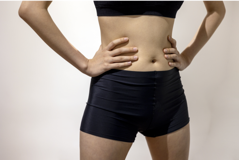 운동용 복장을 한 여성의 복부와 골반 아래를 클로즈업한 사진. 그녀는 검은색 스포츠 브라와 검은색 광택 있는 숏 팬츠를 입고 있으며, 양손을 허리에 얹고 서 있다. 배경은 밝은 색의 단색 벽이다