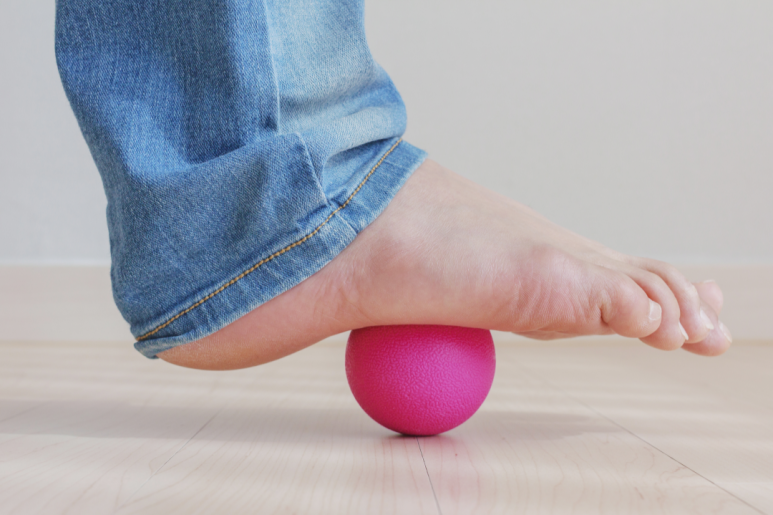 청바지를 입은 사람이 맨발로 바닥 위의 분홍색 공을 밟고 있는 모습. 발바닥 아래의 작은 공을 이용해 발 마사지를 하는 장면으로 보인다