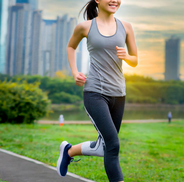 운동복을 입은 여성이 도시 공원 산책로를 따라 달리고 있는 모습. 회색 민소매 상의와 검은색 레깅스를 착용했으며, 배경에는 초록색 잔디와 나무, 그리고 고층 건물이 보인다. 해 질 무렵의 따뜻한 햇살이 장면을 비추고 있다